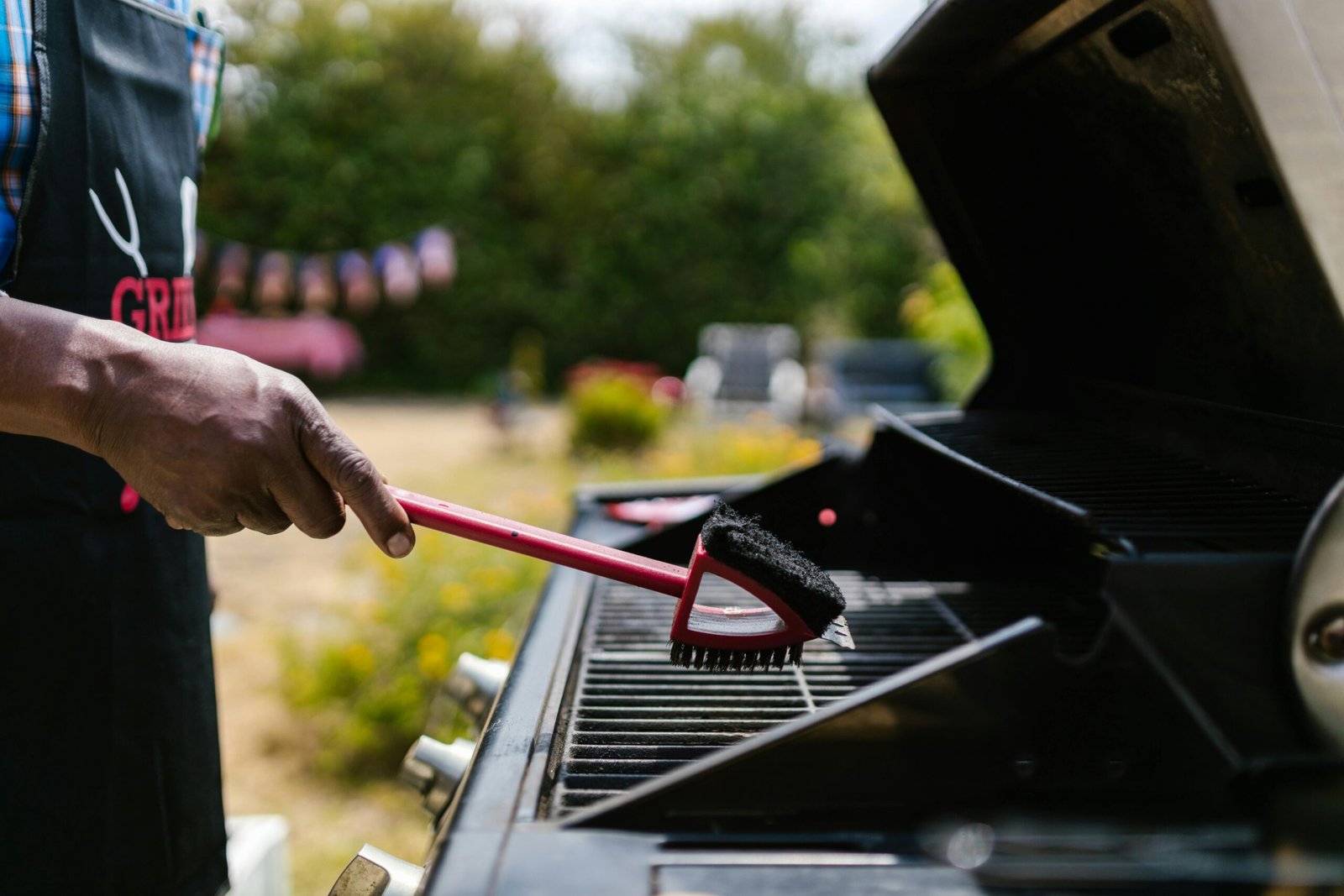 Person cleaning BBQ grill with brush in a sunny backyard setting.